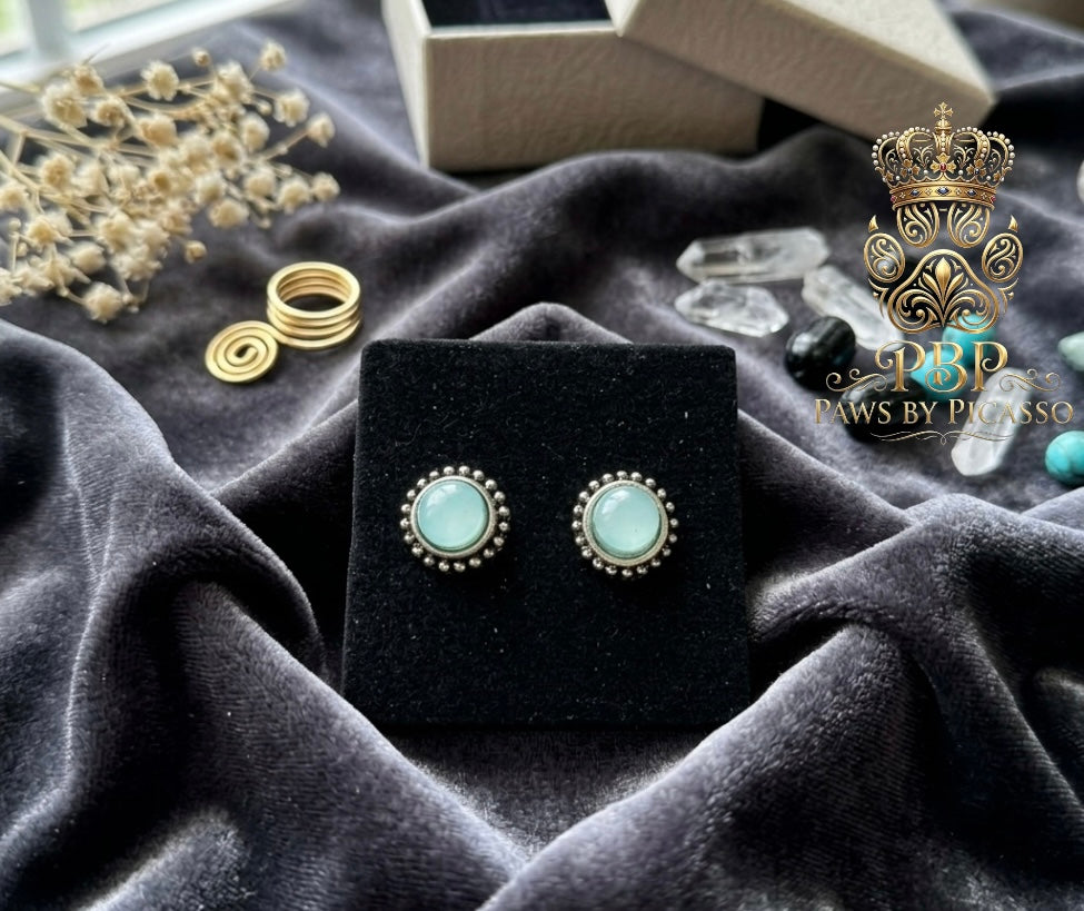 Pair of earrings on a black stand with silver and turquoise stones, surrounded by decorative items on a dark fabric background.