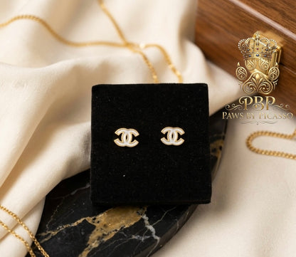 Gold Chanel earrings in a black display on a marble surface with a brand logo in the corner.