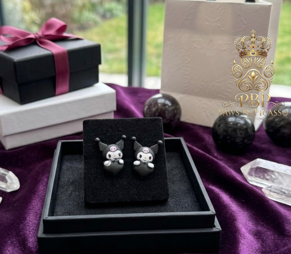 Black cat-shaped earrings on a black stand with a purple ribbon and gift box in the background.