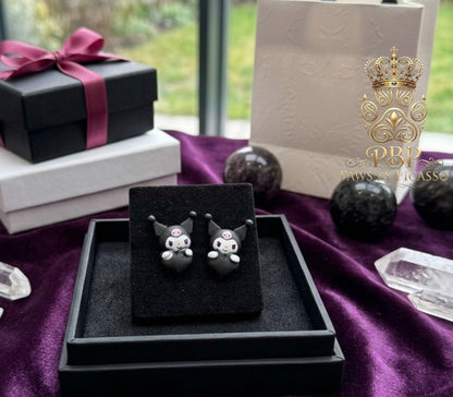 Black cat-shaped earrings on a black stand with a purple ribbon and gift box in the background.