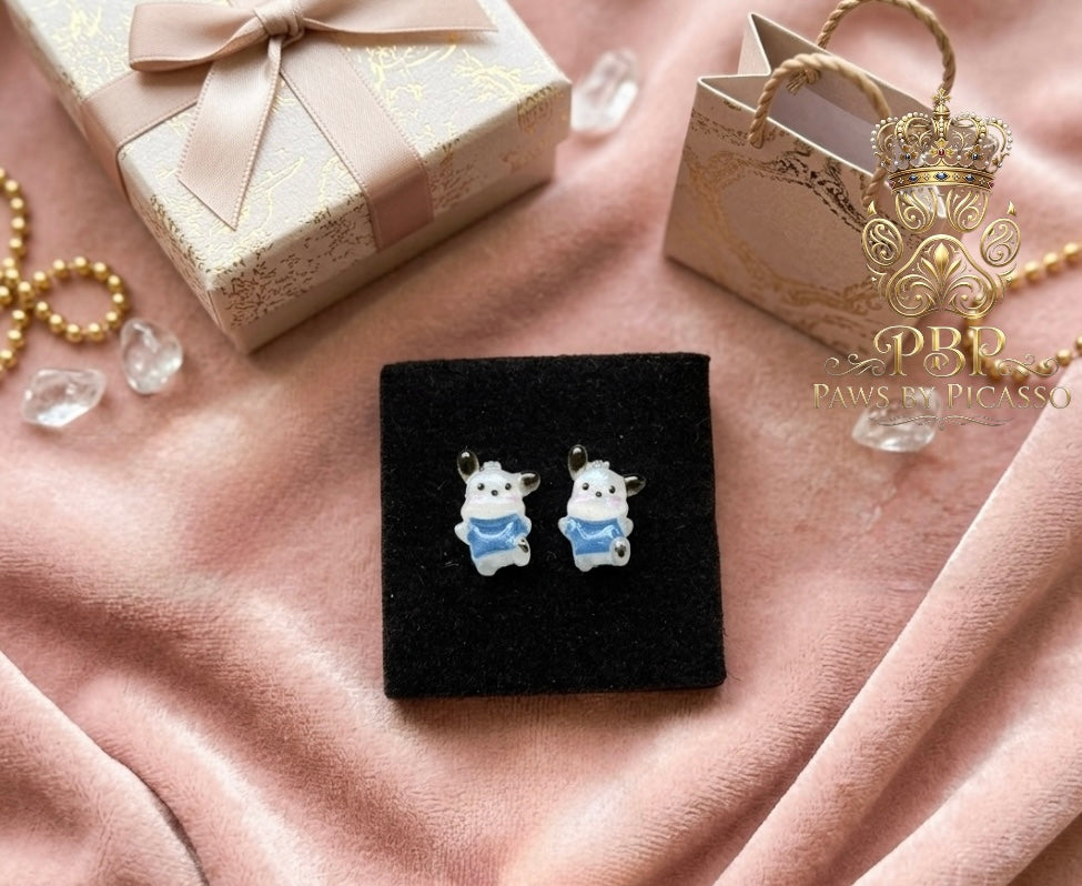 Pair of dog-shaped earrings on a black stand with a gift box and decorative items in the background.