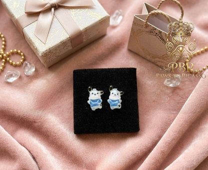 Pair of dog-shaped earrings on a black stand with a gift box and decorative items in the background.