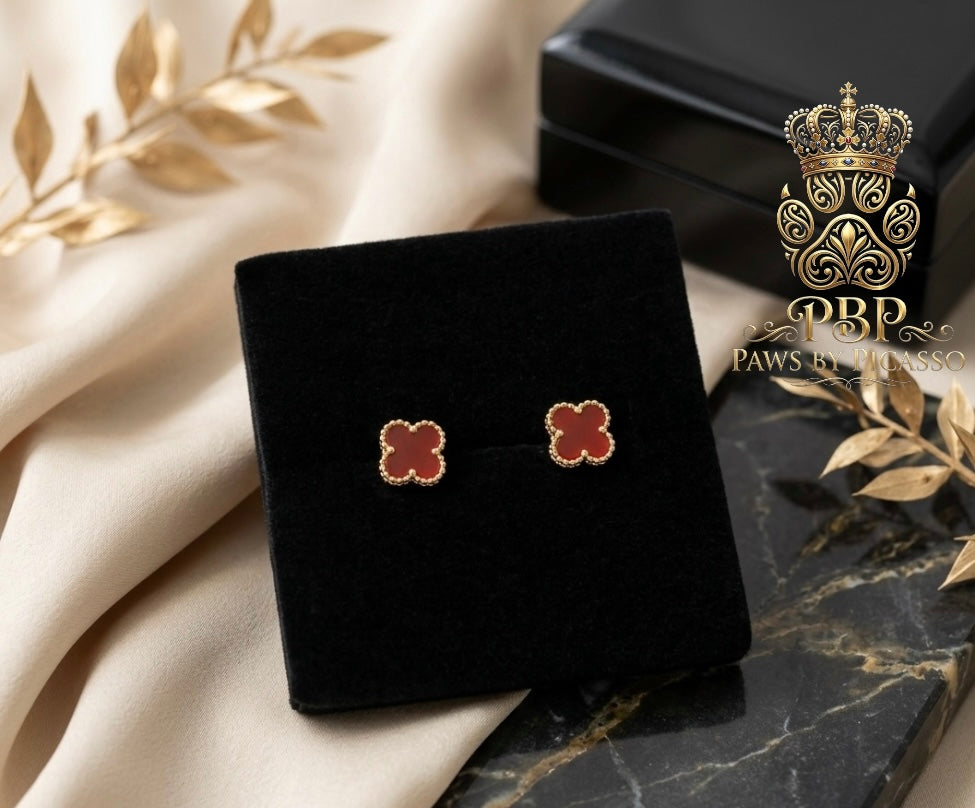 Clover-shaped earrings on a black velvet stand with a decorative box and leaves in the background, featuring the brand 'Paws by Picasso'.