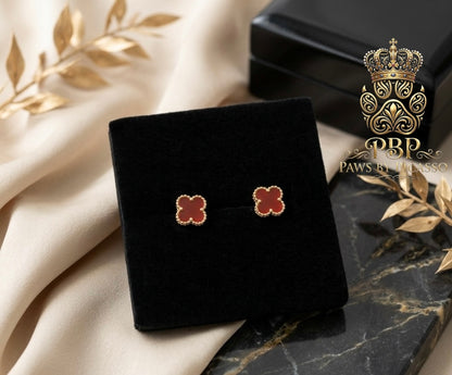 Clover-shaped earrings on a black velvet stand with a decorative box and leaves in the background, featuring the brand 'Paws by Picasso'.