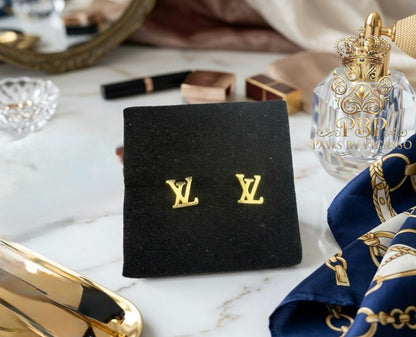 Gold earrings on a black box with a Louis Vuitton logo, next to a perfume bottle and scarf on a marble surface.