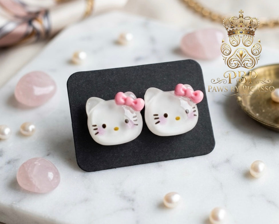 Cat-shaped earrings with pink bows on a black card, surrounded by pearls and gemstones.